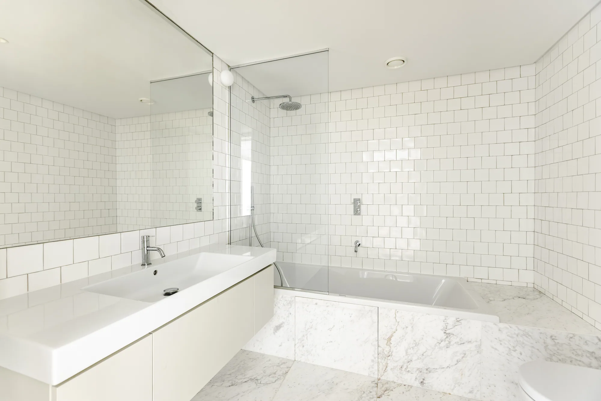 A white-tiled bathroom with a long basin under a wide mirror and a bath behind a glass screen.