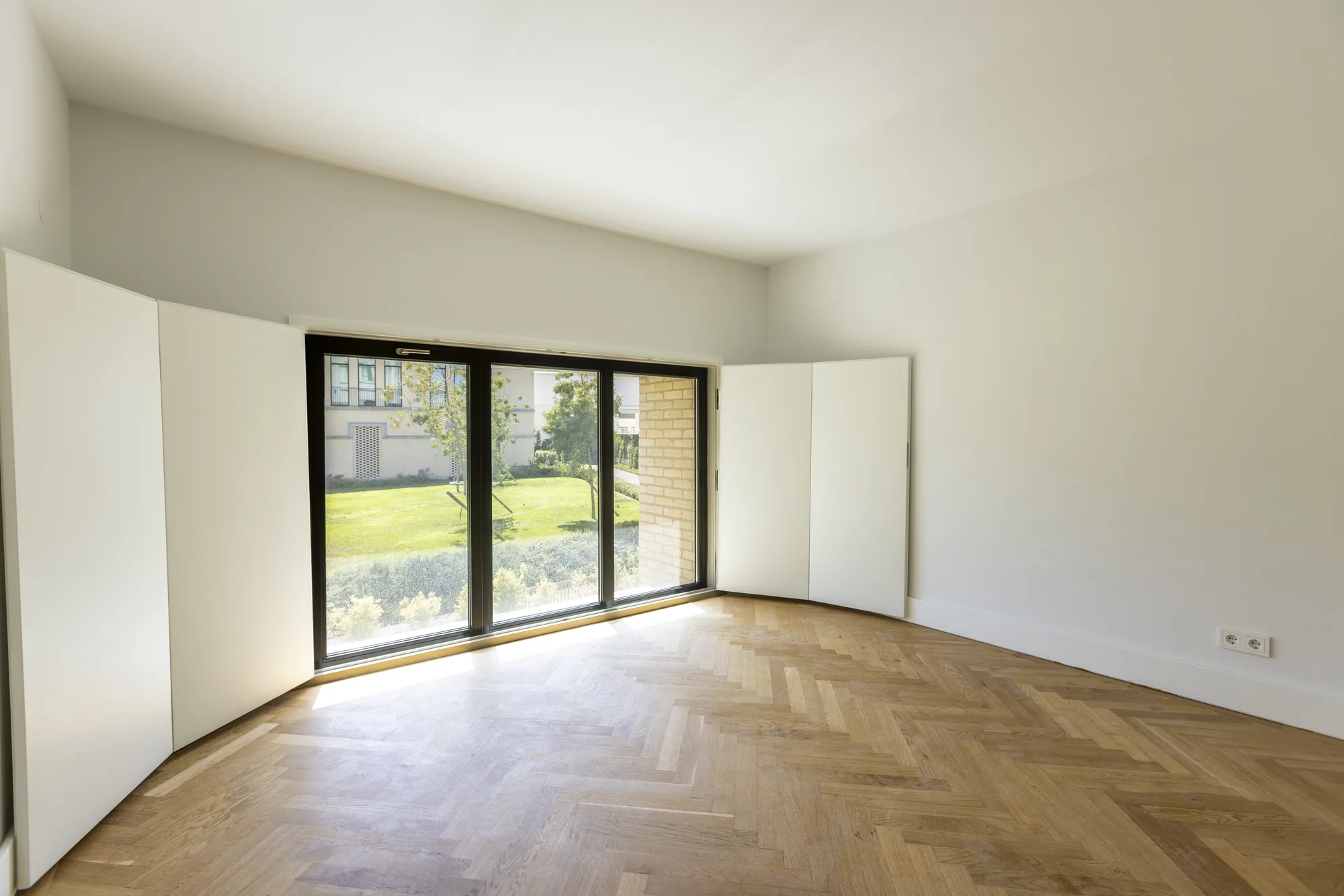 An empty room with a herringbone wood floor and wide glass doors between folded white shutters.