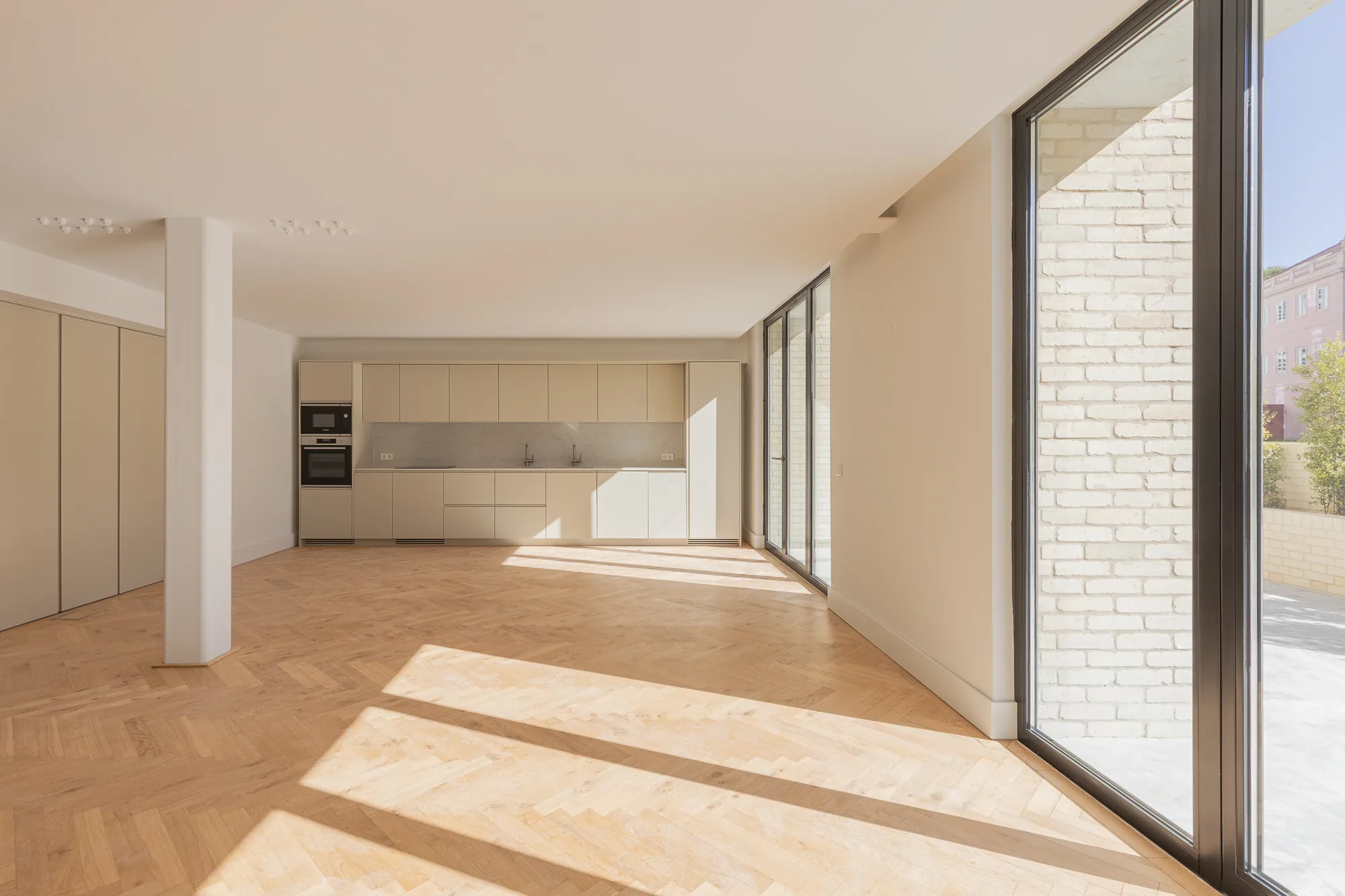 An empty open-plan room with a herringbone wood floor, a white column and a kitchen along the far wall, lit by a tall glass wall on the right.