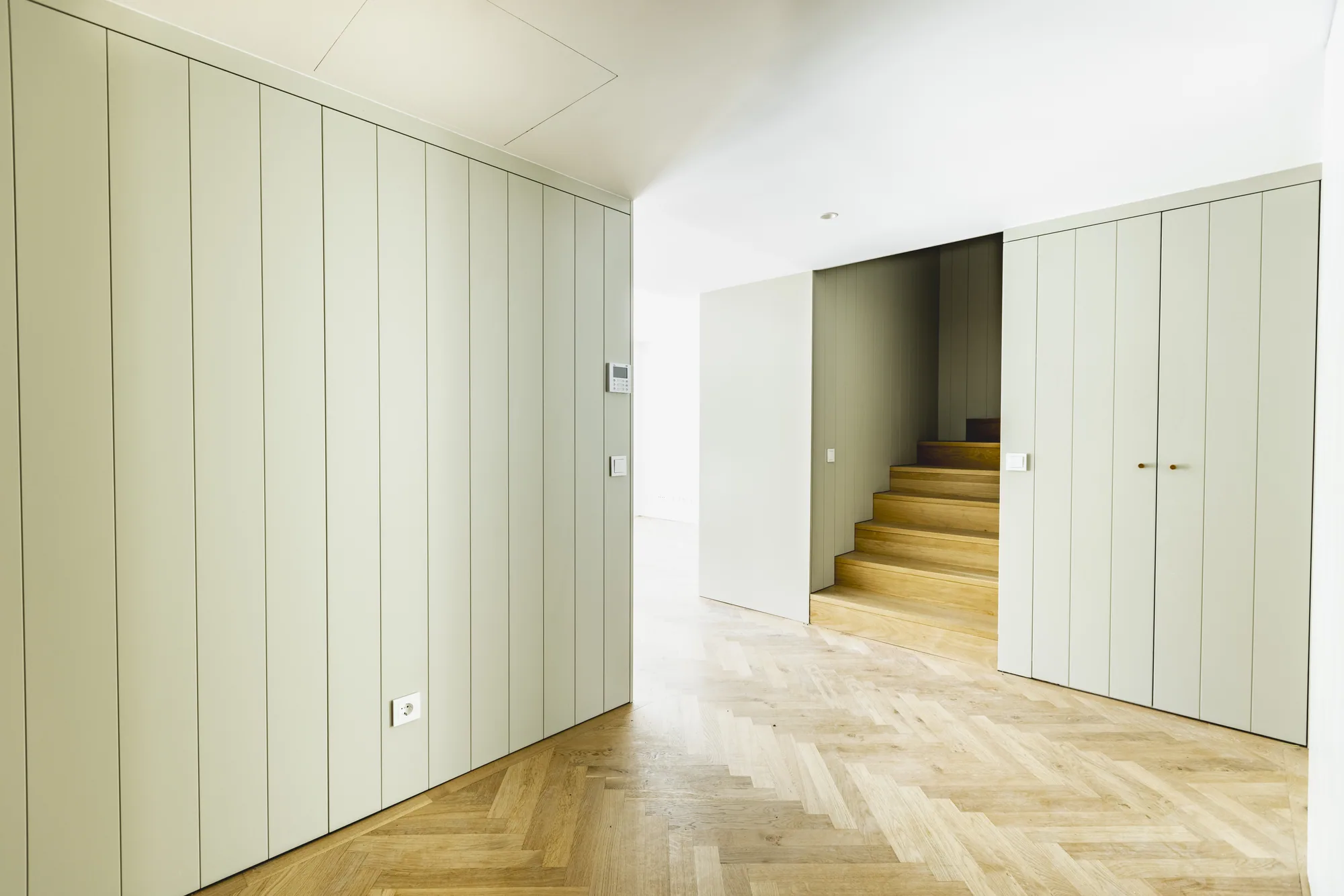 A hall lined with pale panelled cupboards, with a wooden stair rising between them.