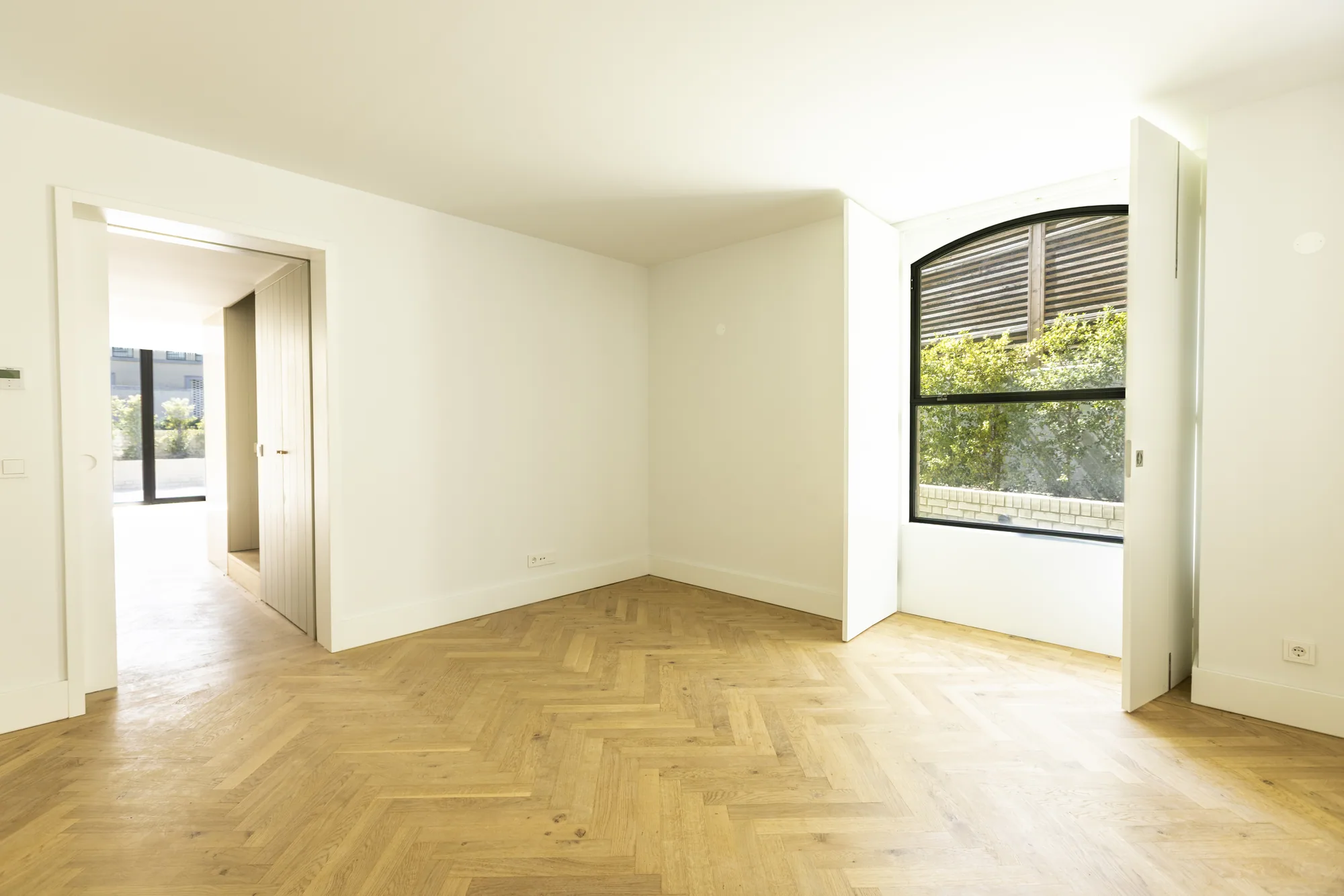 An empty room with a herringbone wood floor and an arched black-framed window between open white shutters, with a doorway through to the living space.