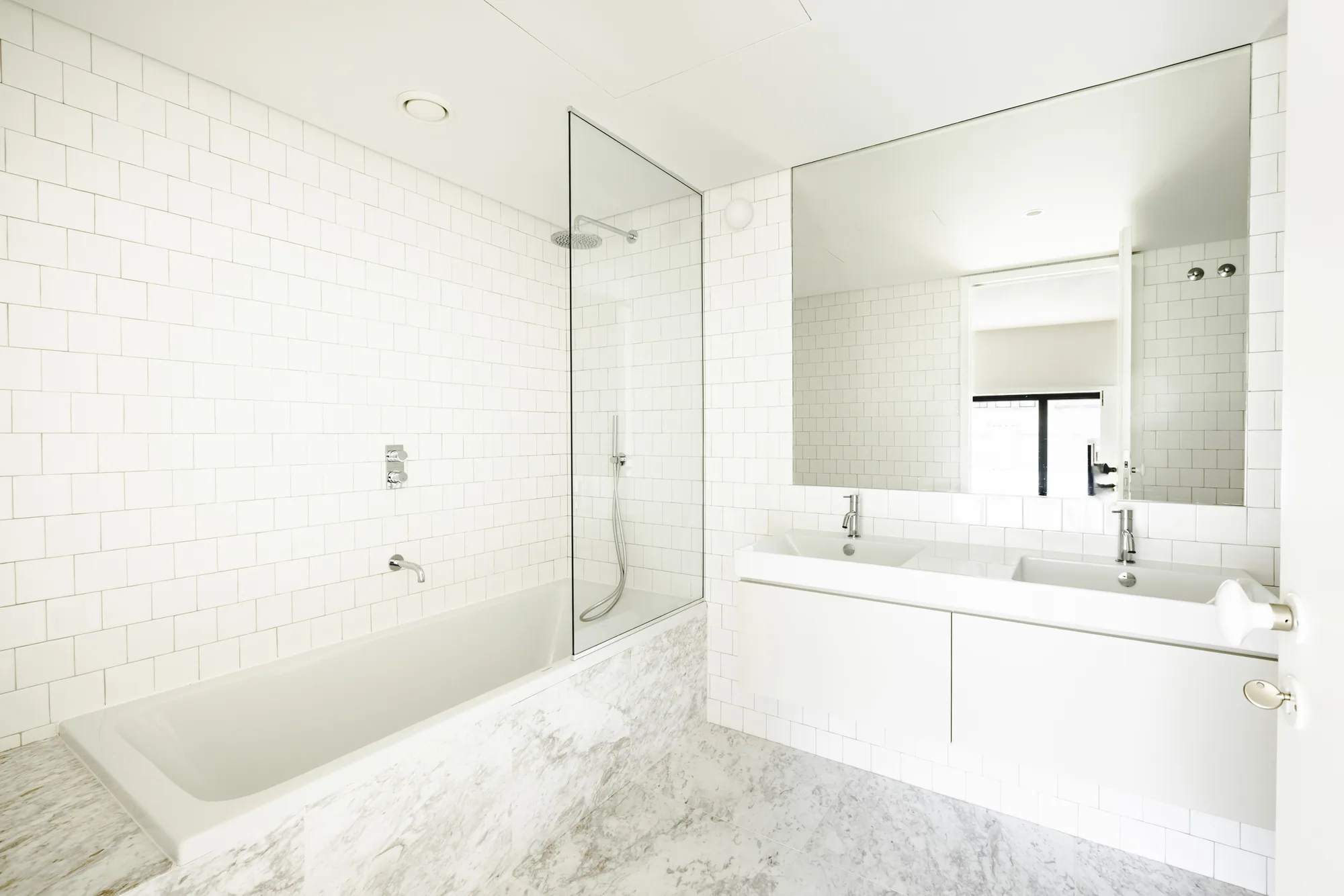 A white-tiled bathroom with a bath behind a glass screen and a long double basin under a wide mirror.