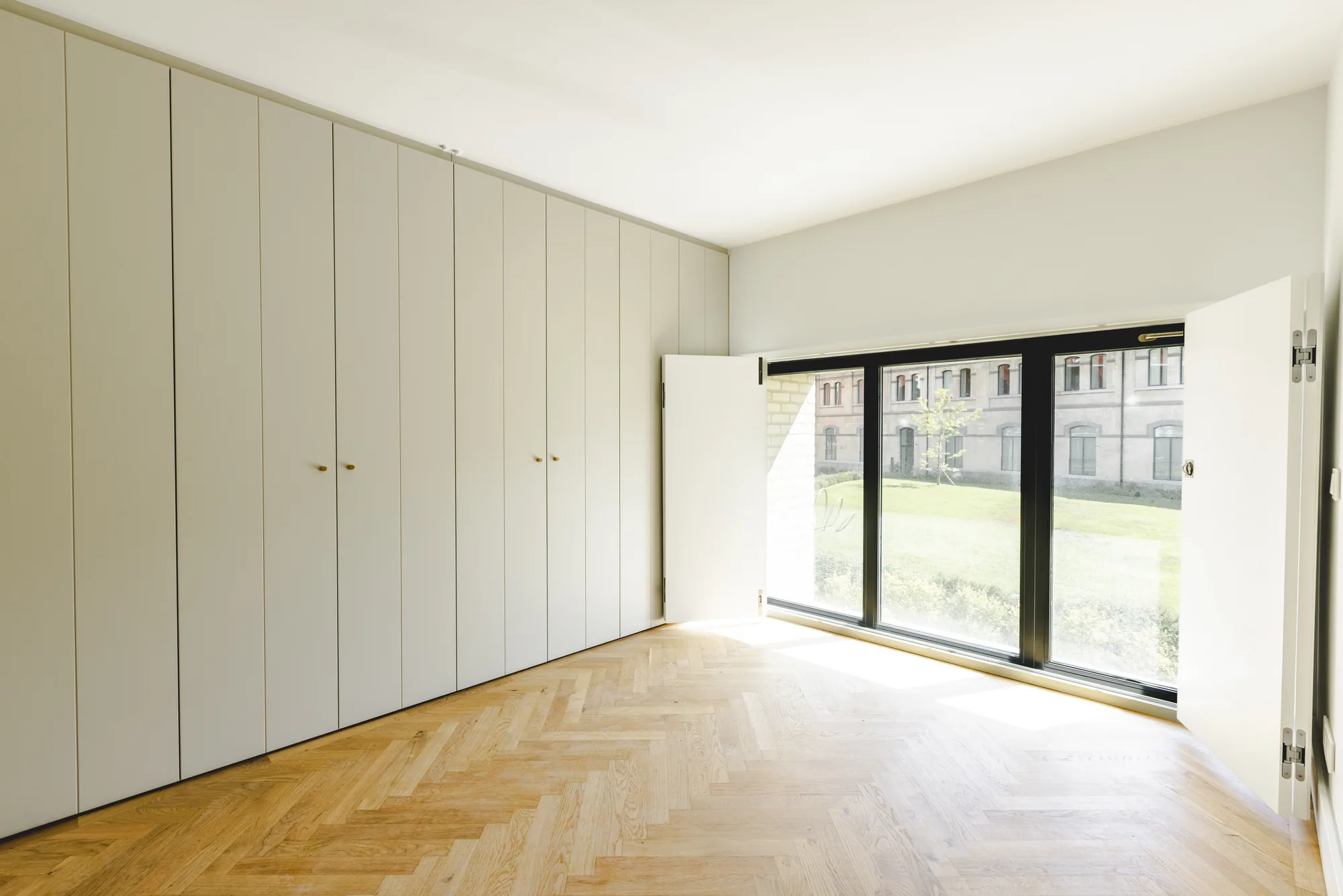 An empty room with a wall of built-in cupboards and a wide window between open white shutters.