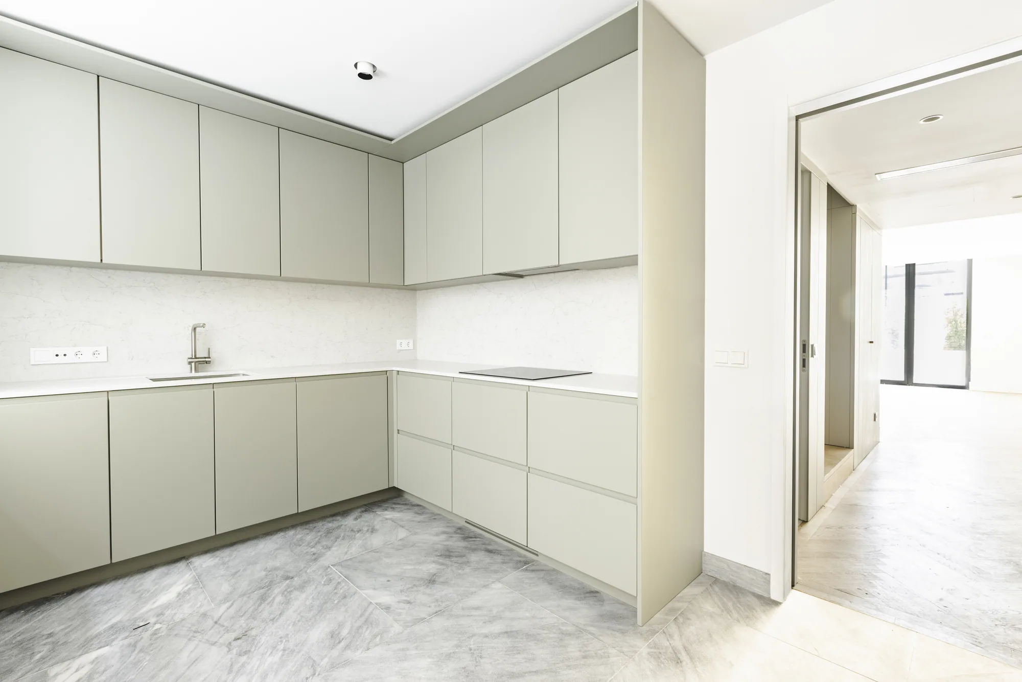An L-shaped kitchen of pale grey-green cabinets over a grey veined floor, with a doorway through to the hall.