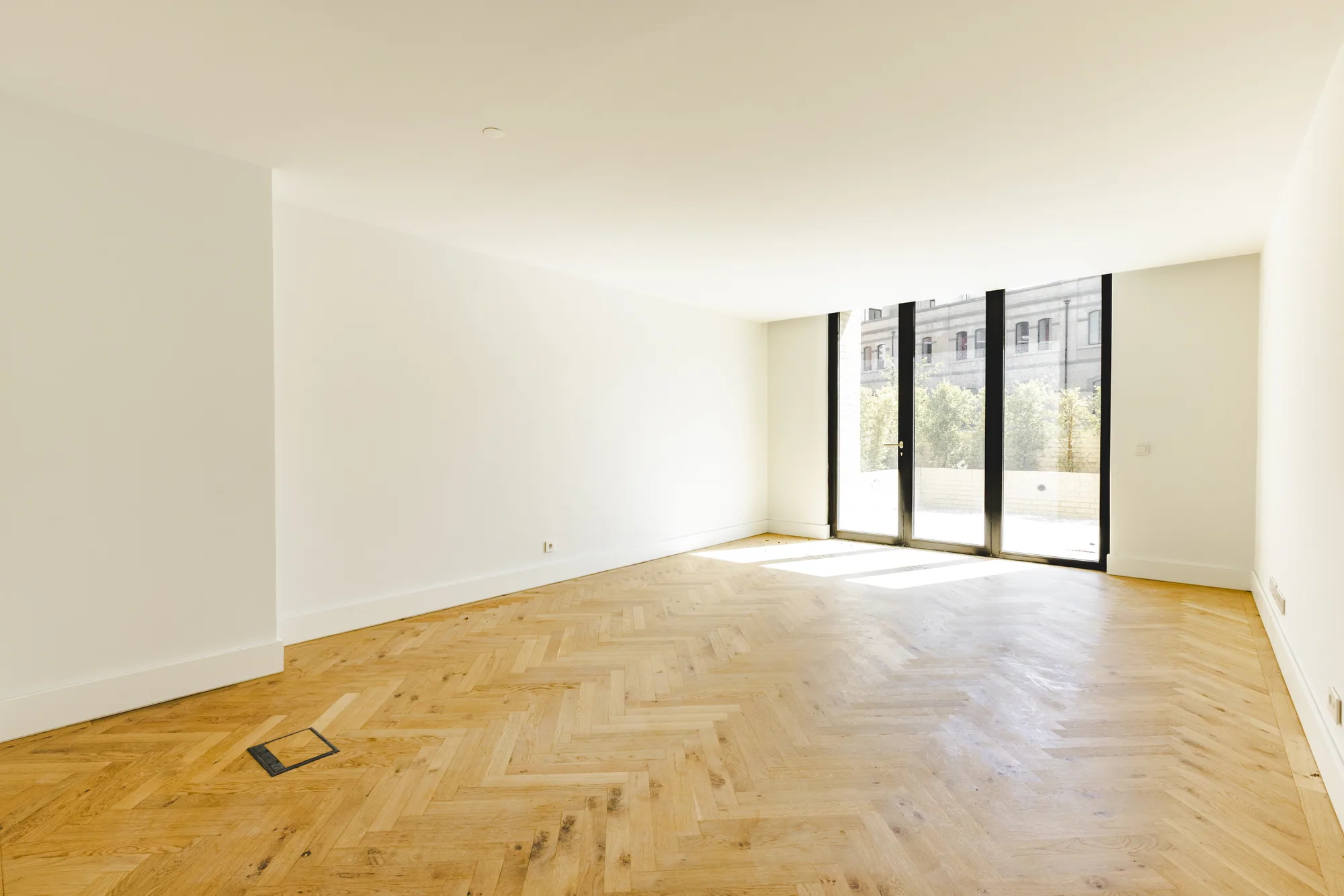 An empty room with a herringbone wood floor and tall glass doors at the far end letting in sunlight.