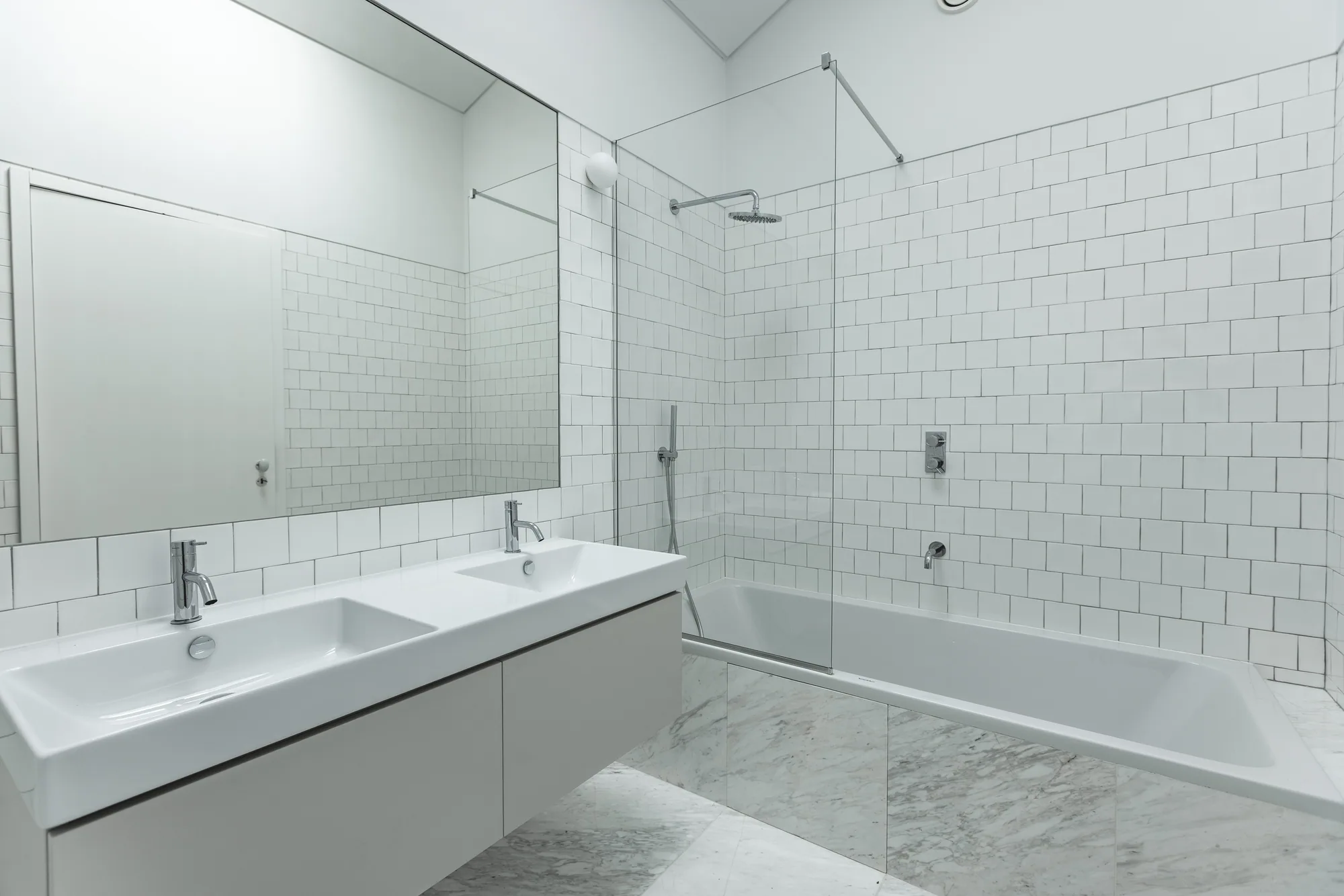 A white-tiled bathroom with a double basin under a wide mirror and a bath behind a glass screen.