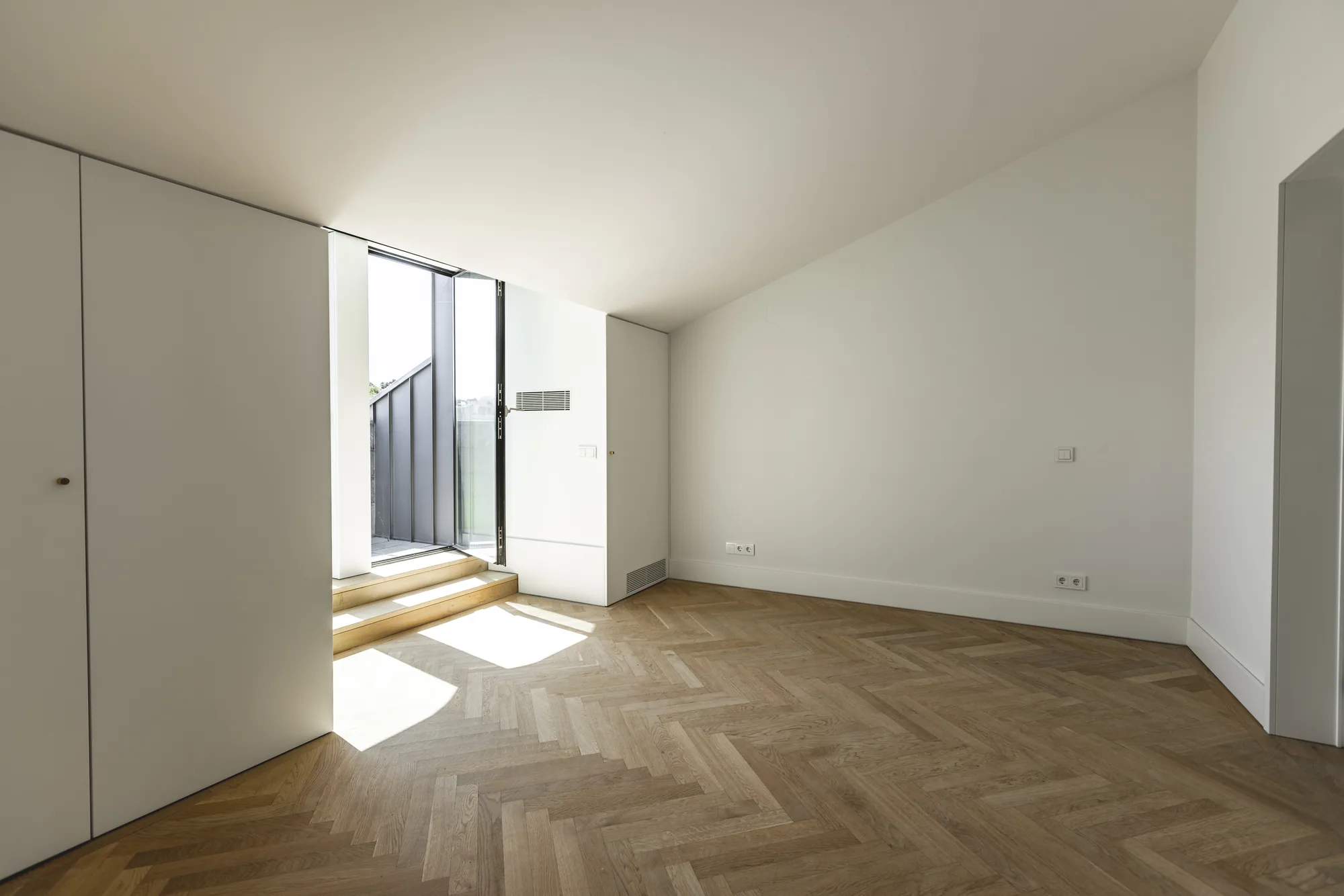 An empty room under a sloping ceiling with a tall white cupboard, a herringbone wood floor and wooden steps up to an open glass door.