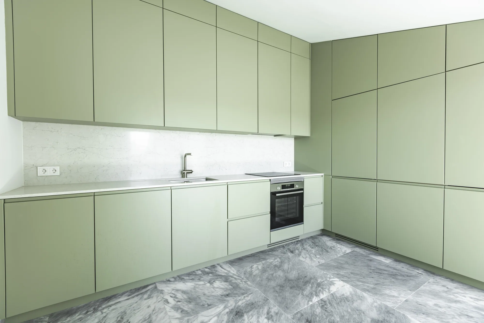 A kitchen fitted with sage-green cabinets on two walls, with an oven, a sink and large grey floor tiles.