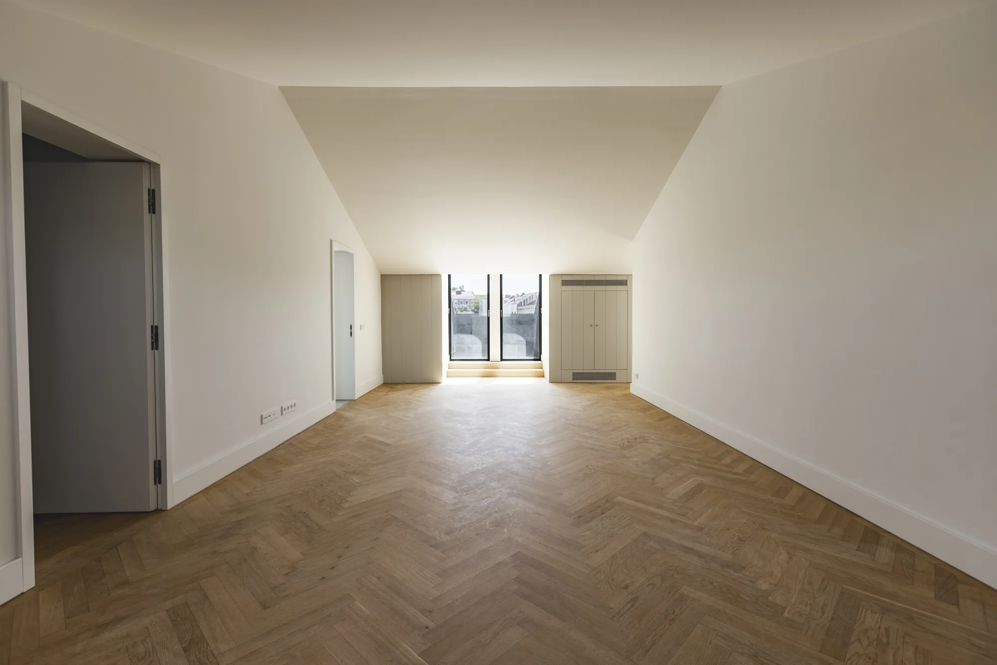 A long empty room under a sloping ceiling, with a herringbone wood floor and a pair of tall windows at the far end.