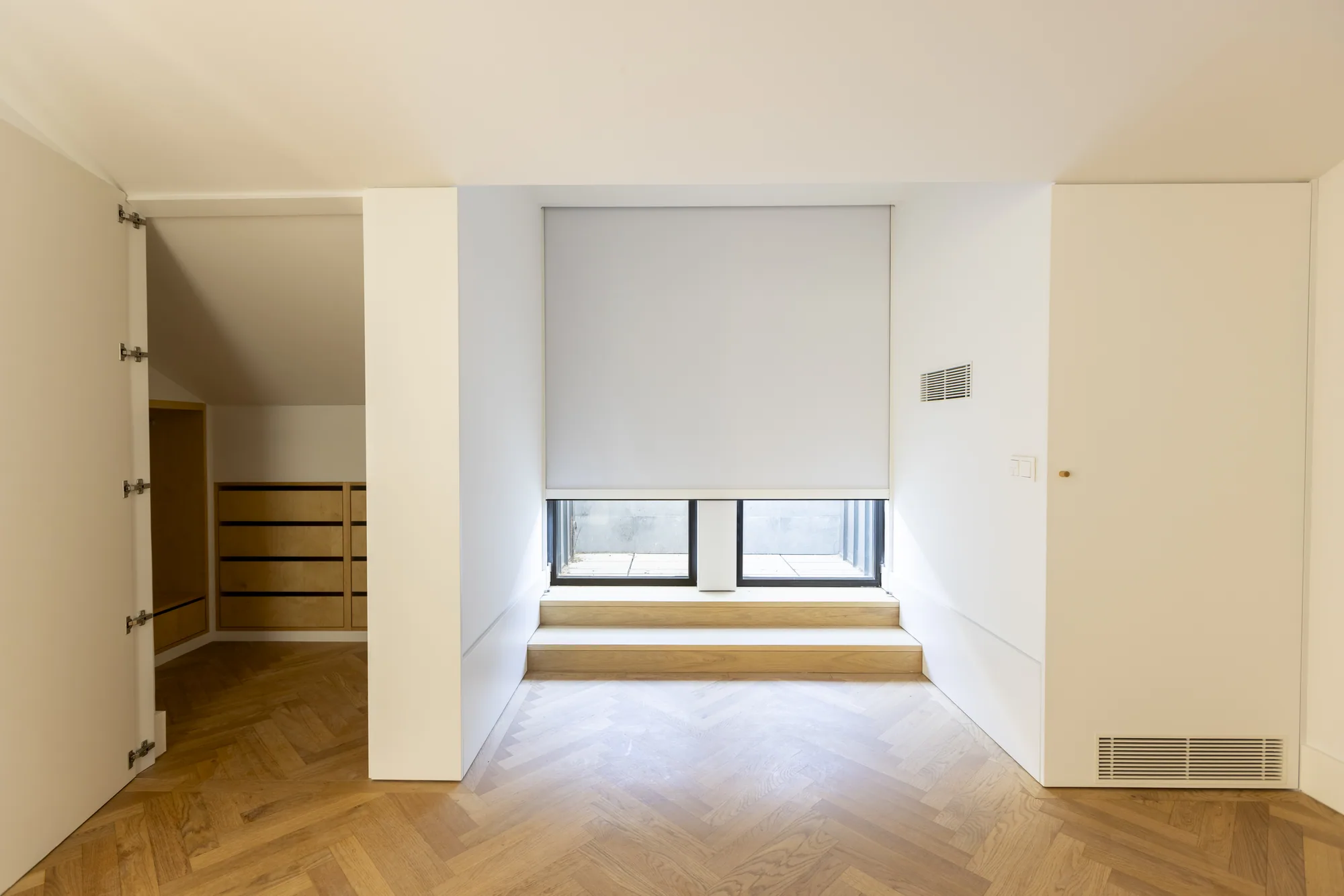 An empty room with a herringbone wood floor, a low window under a half-lowered blind above two wooden steps, and an open cupboard with drawers on the left.