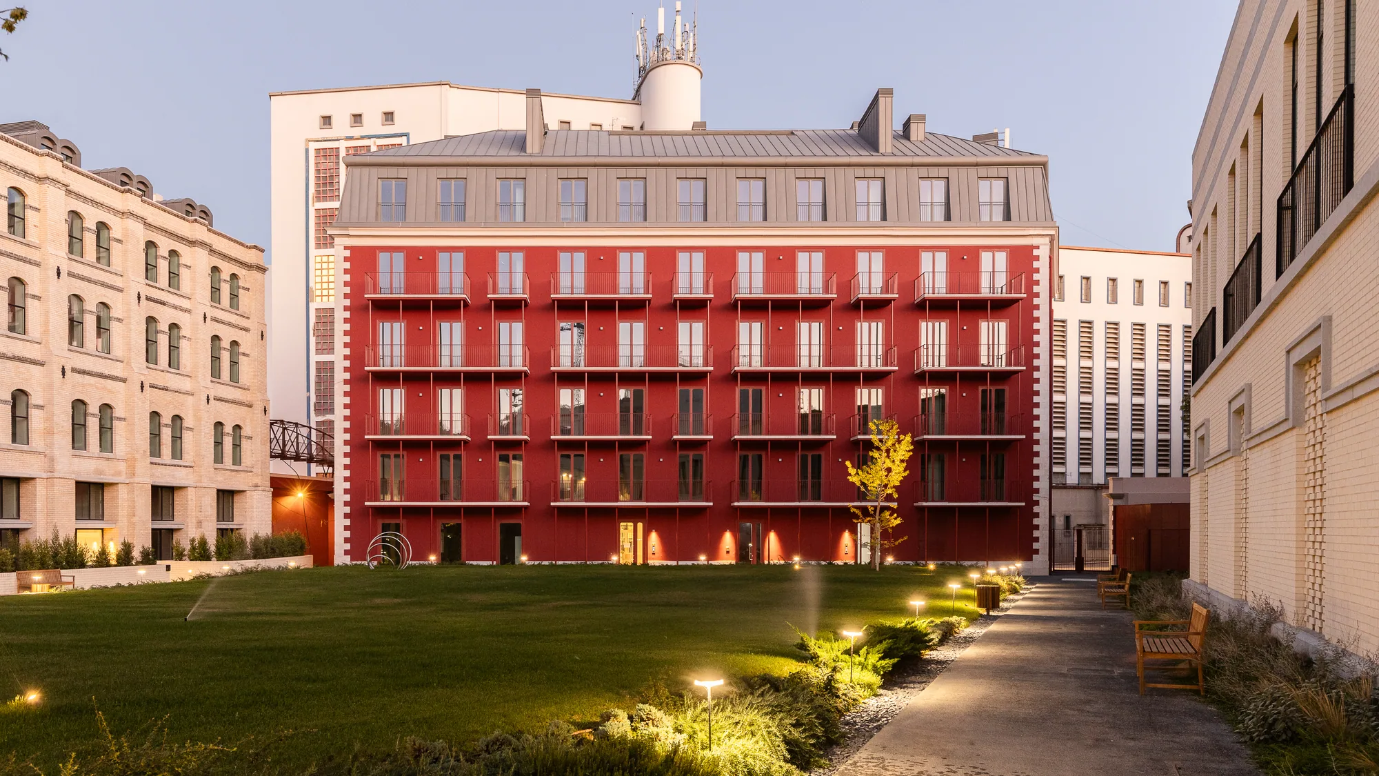 The courtyard at dusk: low lamps along a path through the lawn toward the red building with its lit balconies.