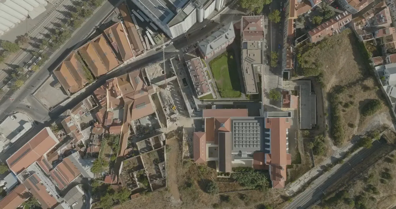 Looking straight down from a drone as it slowly descends and turns over the historic block: tiled roofs, a square roof of skylights and a green lawn.