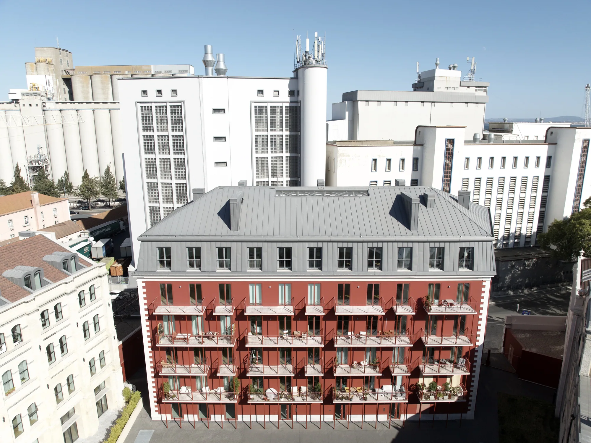 The red building with white stone frames and a grey mansard roof, seen from the air, with tall white silos and industrial buildings behind it.