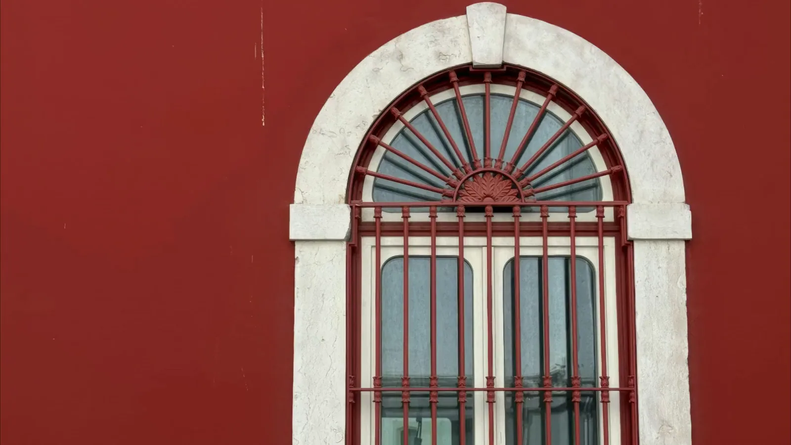 An arched window in a white stone frame with a red iron fan grille, in the red façade on Rua do Beato.