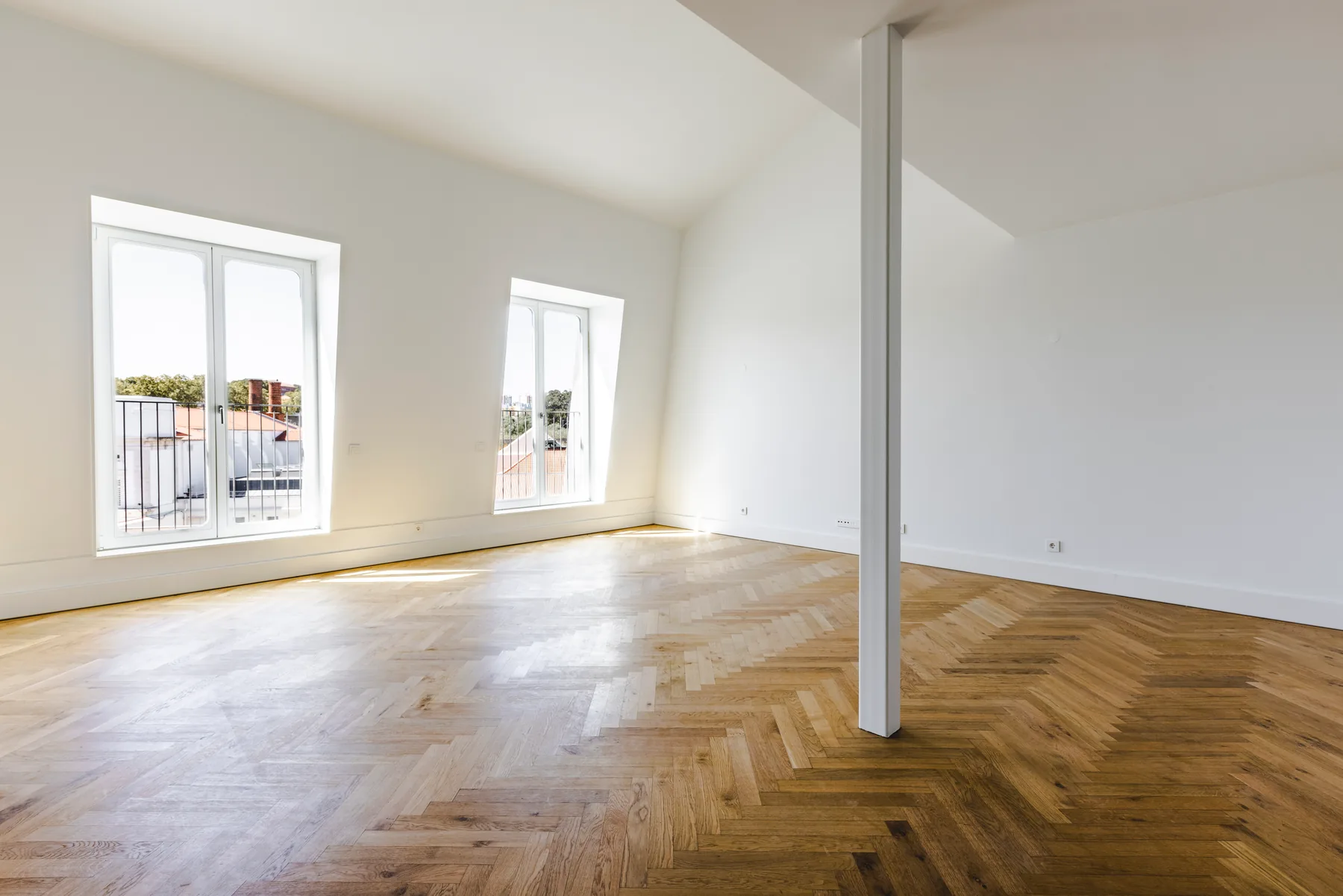 The empty living room: a slender white column, a herringbone wood floor and two tall white-framed windows.
