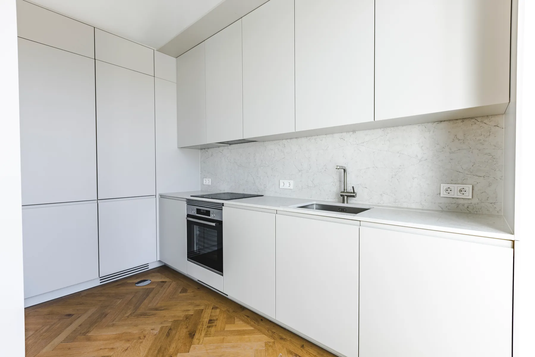 A white kitchen with a pale veined backsplash, a built-in oven and tall cupboards, on a herringbone wood floor.