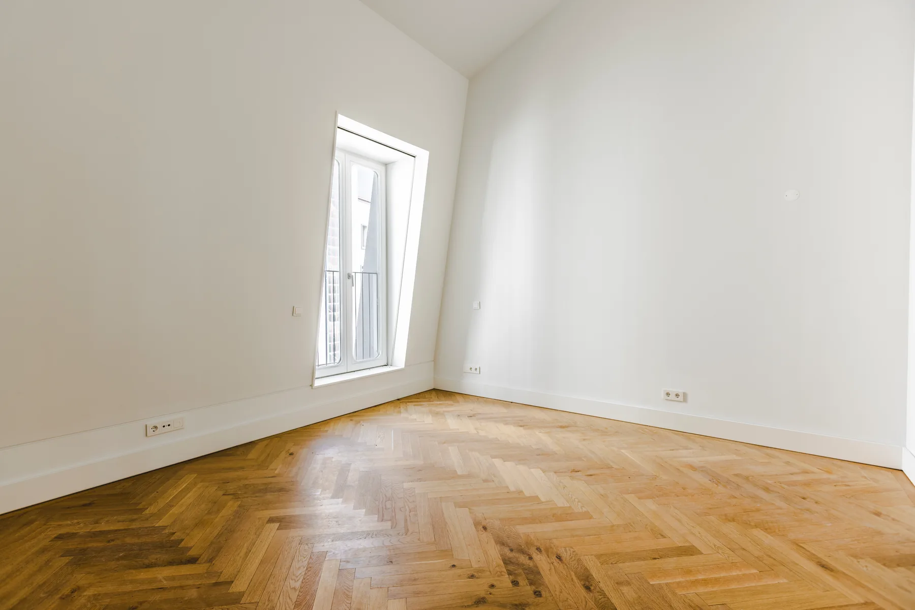 An empty bedroom with a herringbone wood floor and one tall French window set into the sloping wall.