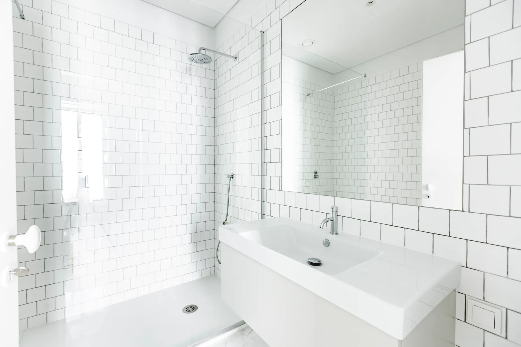A white-tiled bathroom with a glass shower, a wide basin and a mirror.