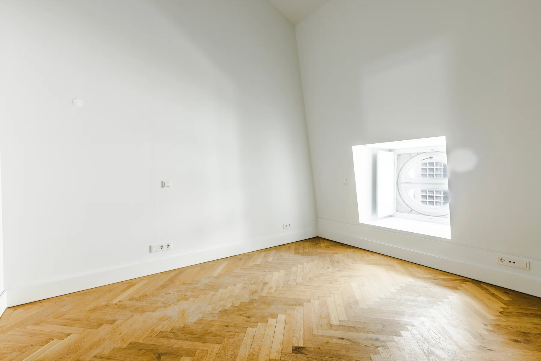 An empty bedroom with a herringbone wood floor and an oval window set deep in the wall.
