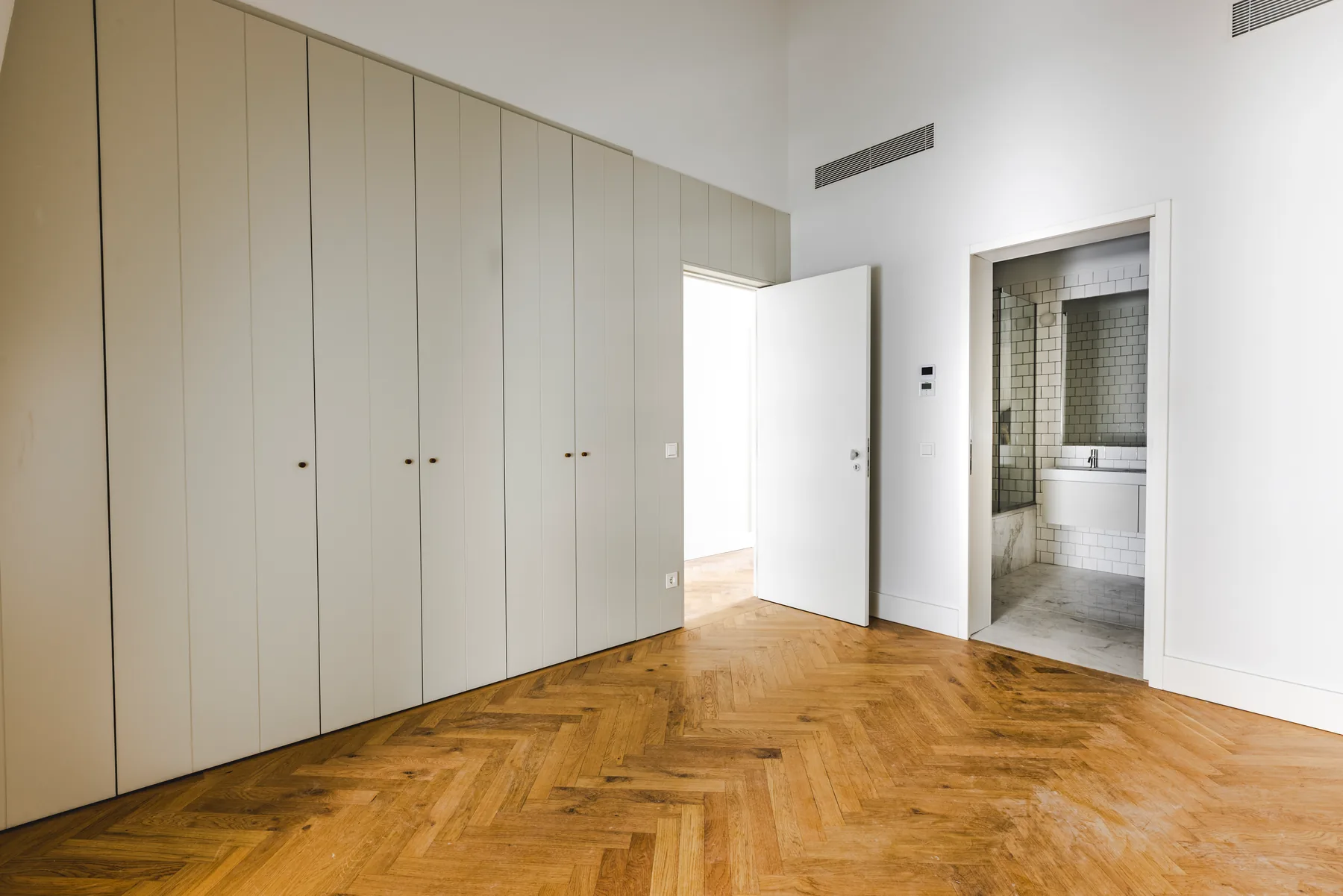 The same bedroom towards the doors: a wall of built-in wardrobes, the open door to the corridor and a bathroom with a bath beside it.