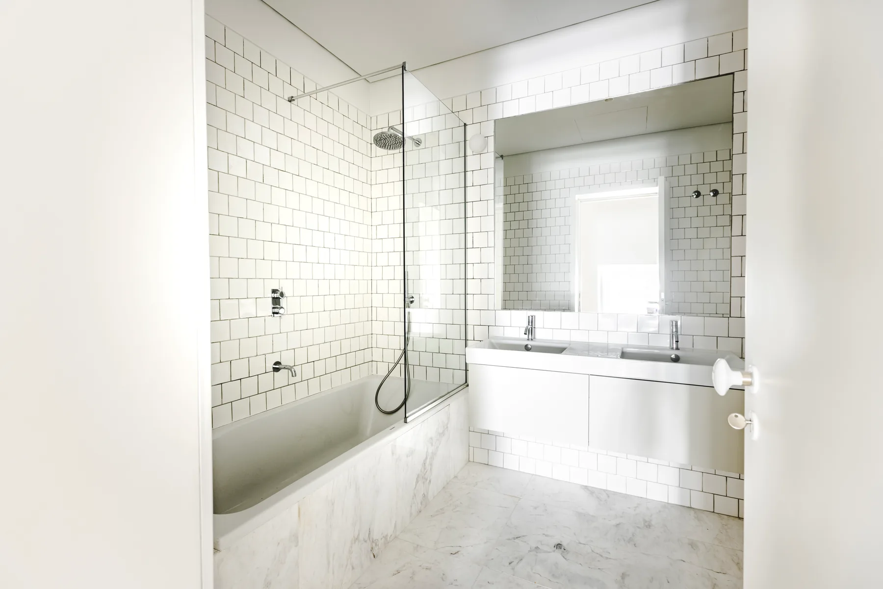 A white-tiled bathroom with a bath behind a glass screen and a double basin under a wide mirror.