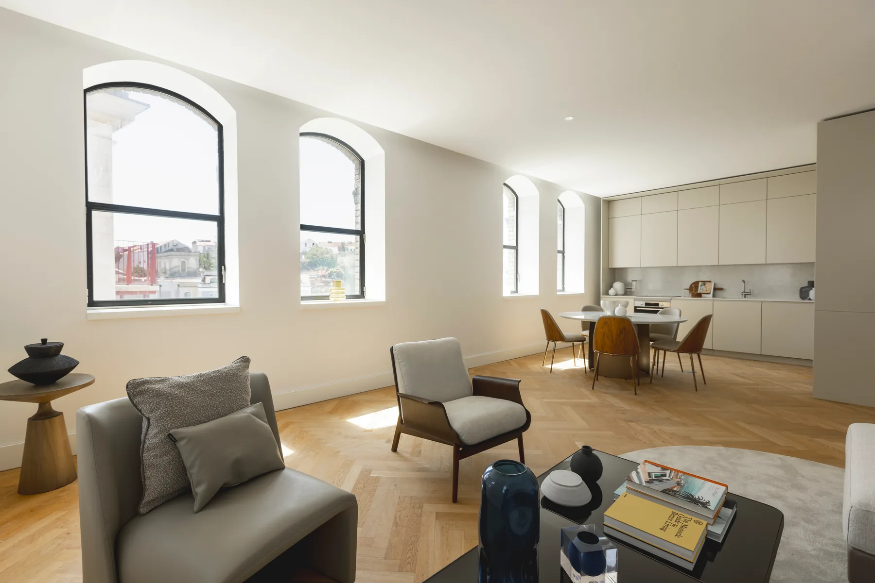 The staged living and dining room seen from the sofa: arched windows along the left wall, a round dining table and the kitchen units along the far wall.