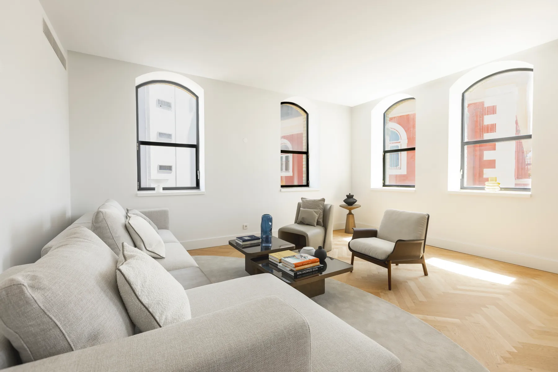 A corner of the living room with a pale sofa, a low table and two armchairs, under four arched windows that look onto a red building opposite.