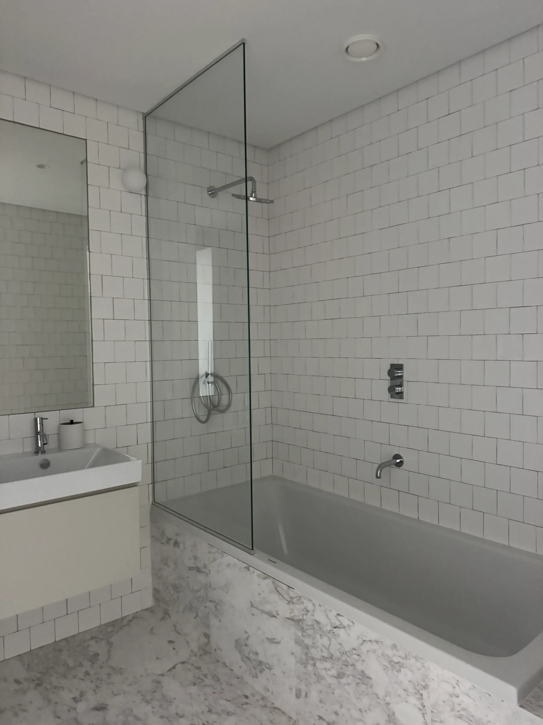 A white-tiled bathroom with a bath behind a glass screen and a basin with a mirror.