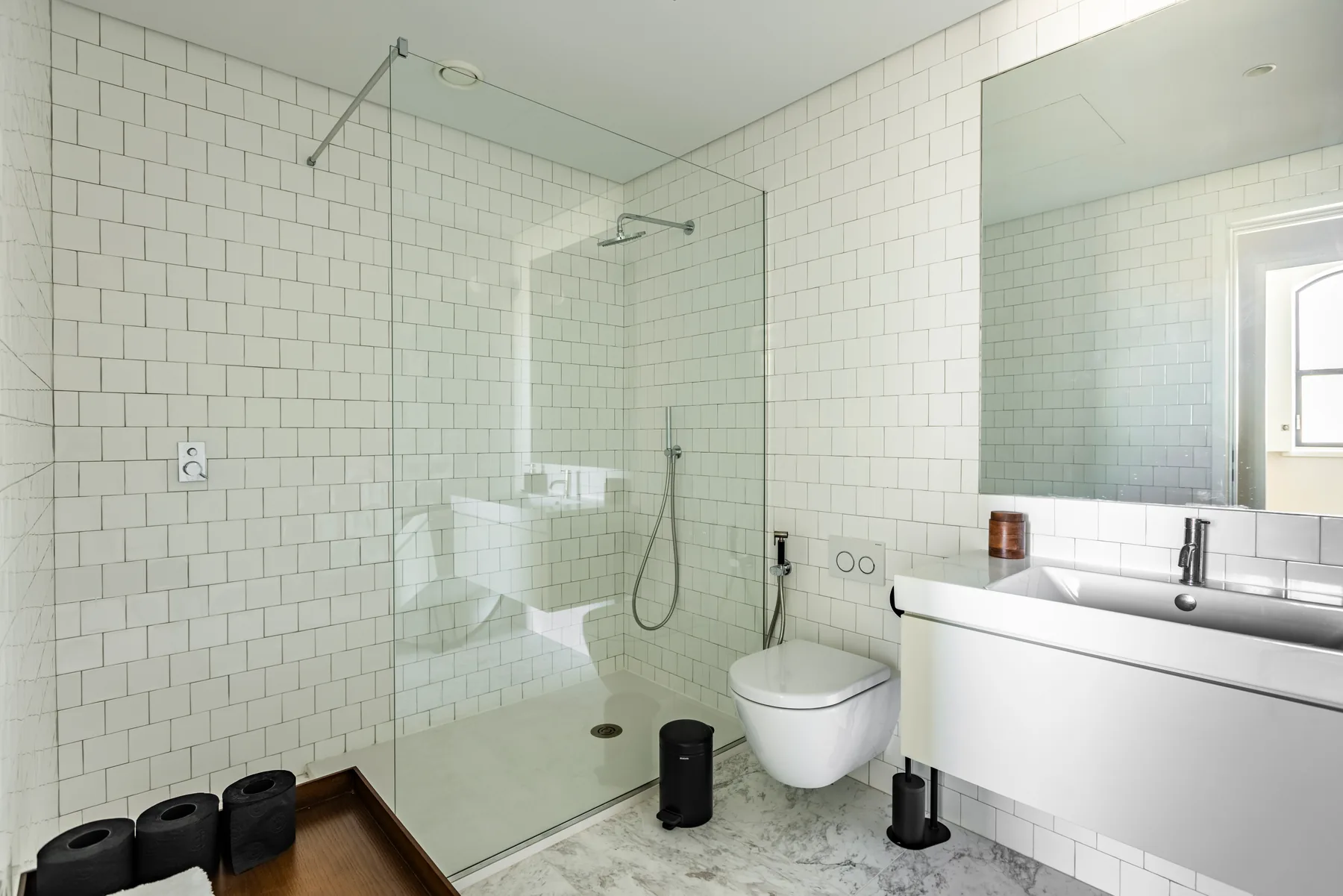 A white-tiled bathroom with a glass walk-in shower, a wall-hung toilet and a long basin under a mirror.