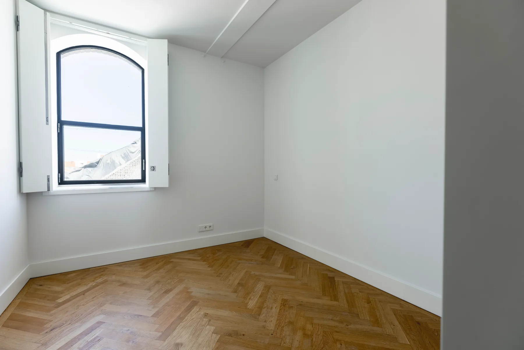 An empty bedroom with a herringbone wood floor and an arched window with folding shutters near the corner.