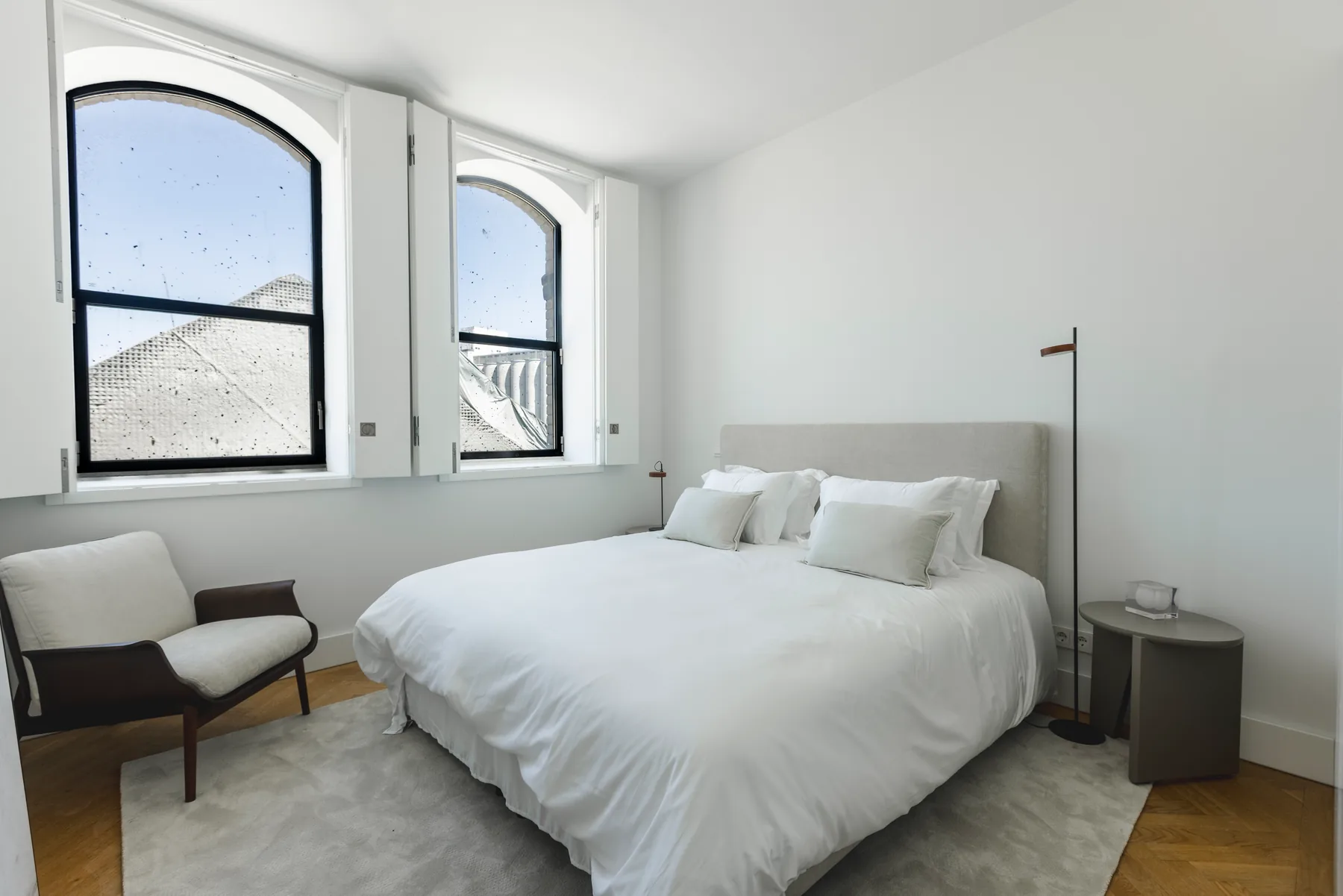 The staged bedroom with a white double bed, an armchair and two arched windows.