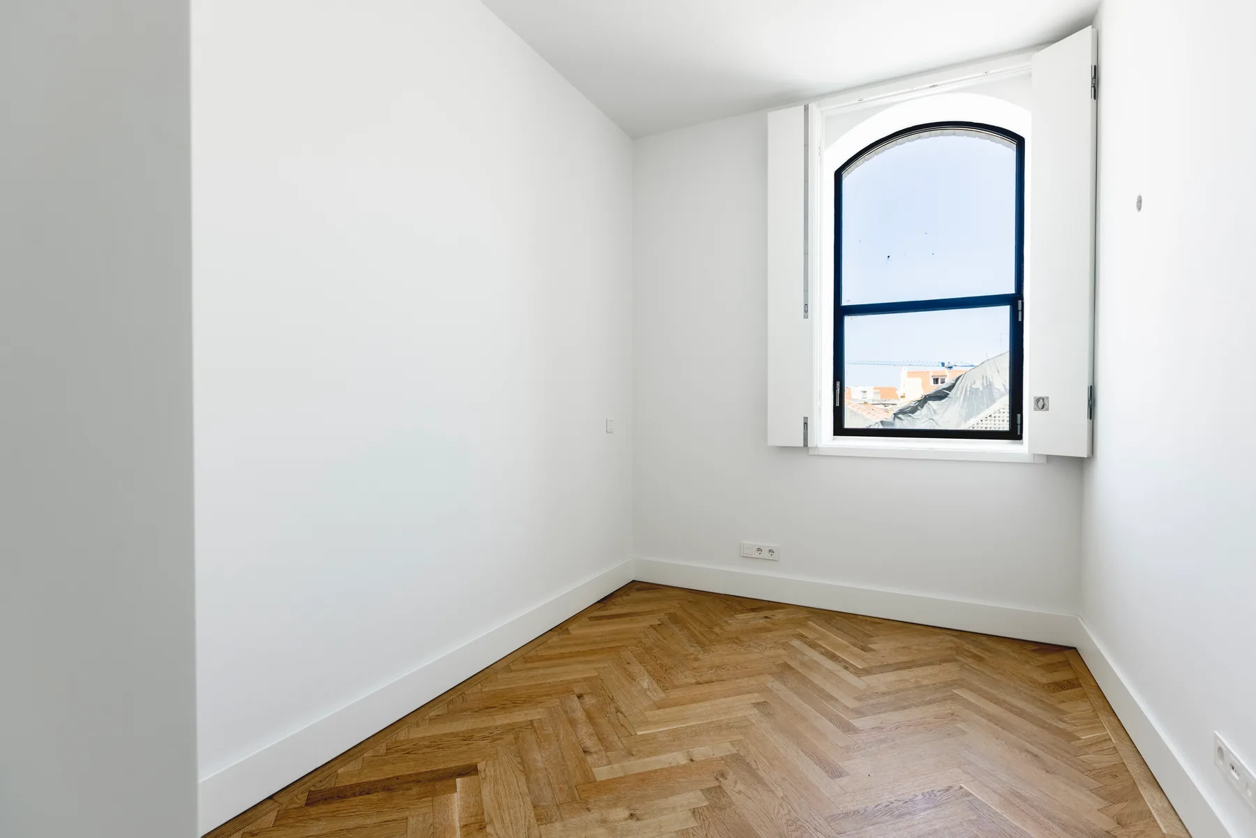 An empty bedroom with a herringbone wood floor and one arched window with folding shutters.